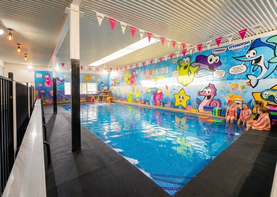 The AQUAKids indoor teaching pool, with the painted mascot mural along the far wall and children waiting at the poolside in towels.